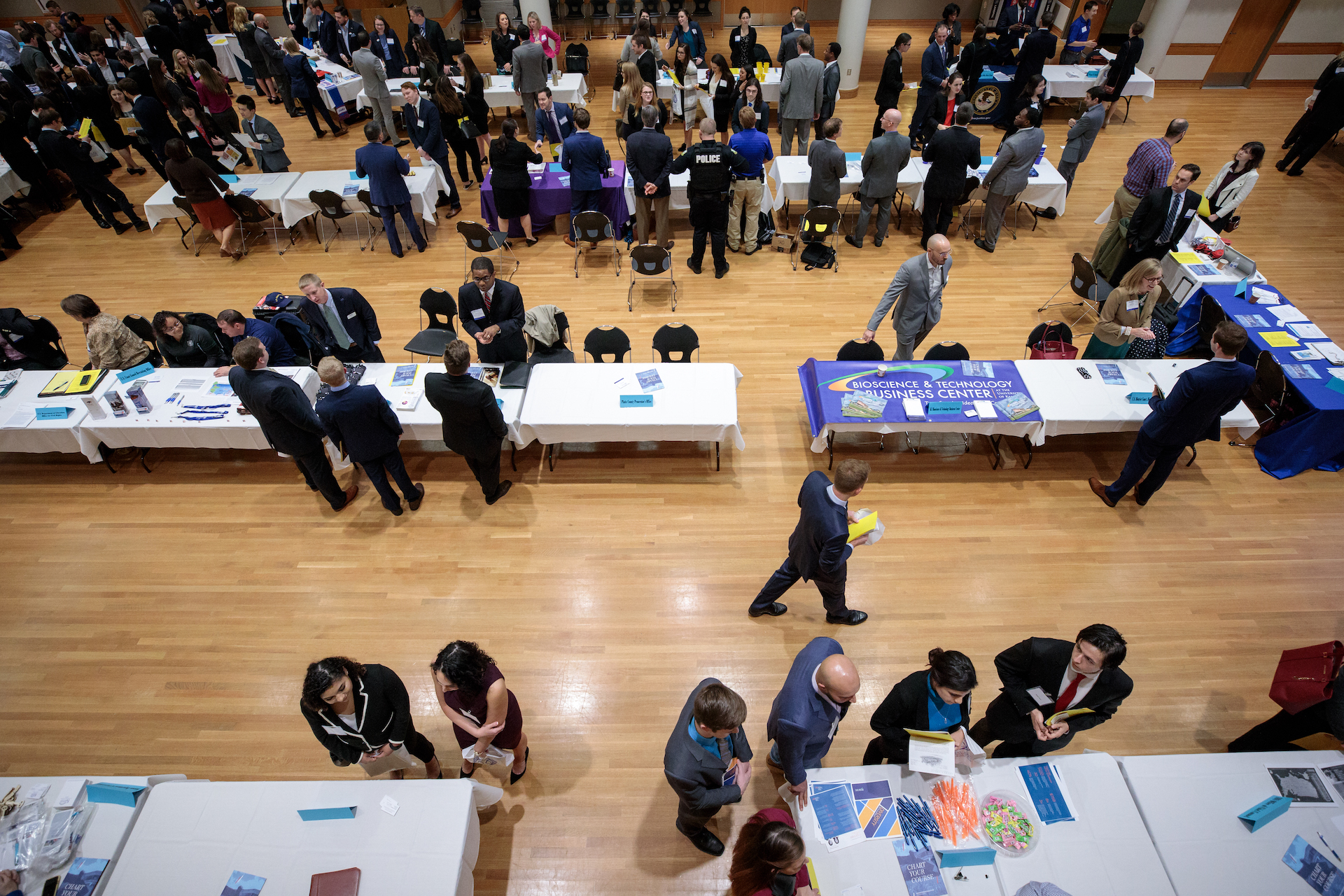 Viewed from overhead, a large number of people in professional attire walk and talk amongst tables filled with business materials.