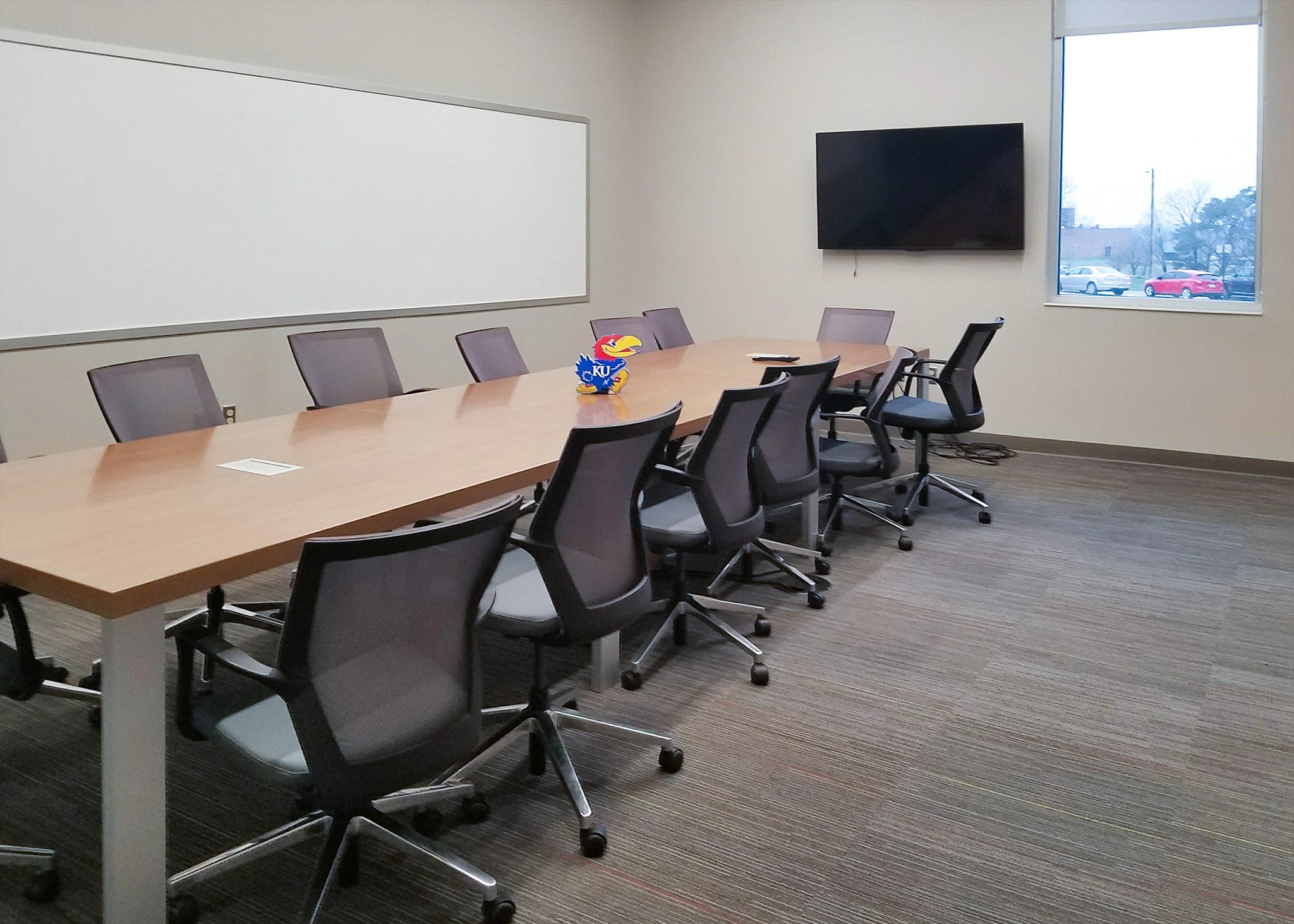 Chairs line a long conference table on two sides; the walls have a whiteboard and a video monitor.