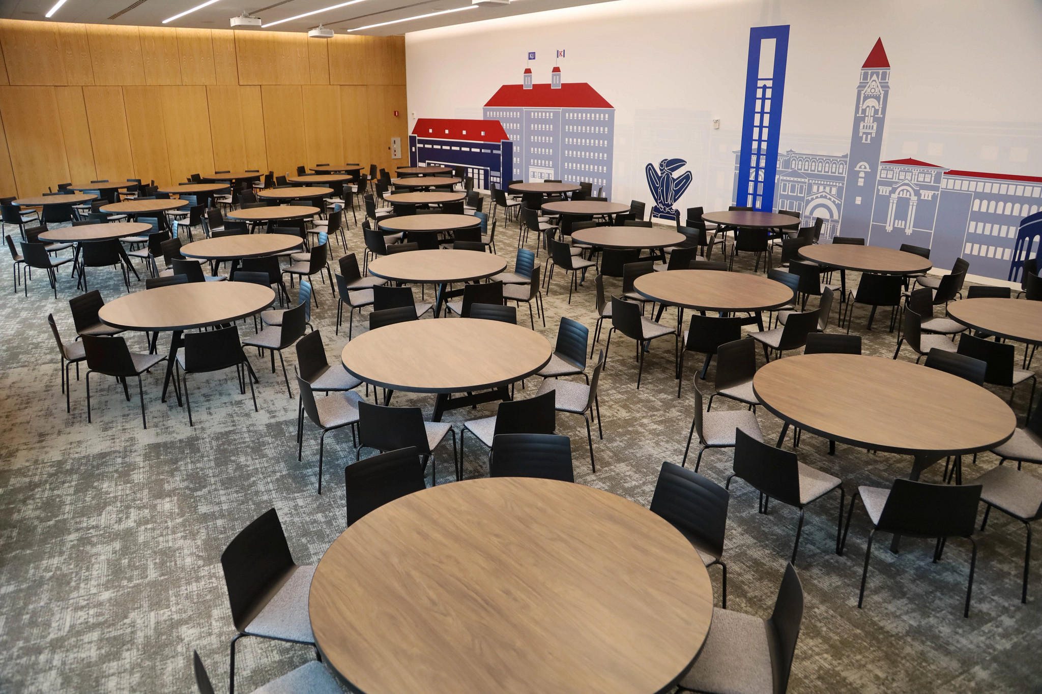 Rows of round table surrounded by chairs, with a KU campus mural on the wall.