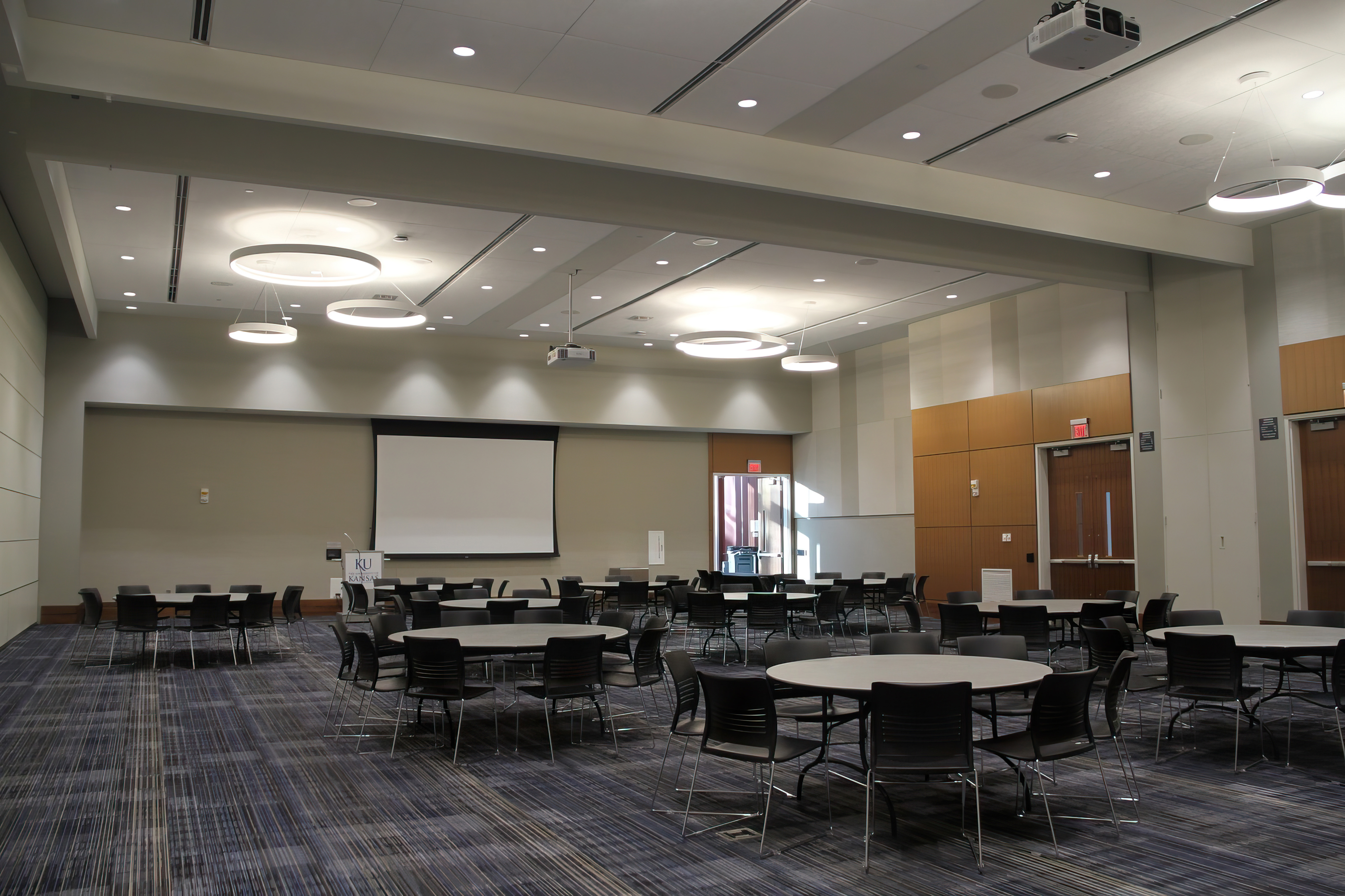A conference room, Forum AB, filled with round tables and chairs, a projector screen at the front, and hanging circular lighting