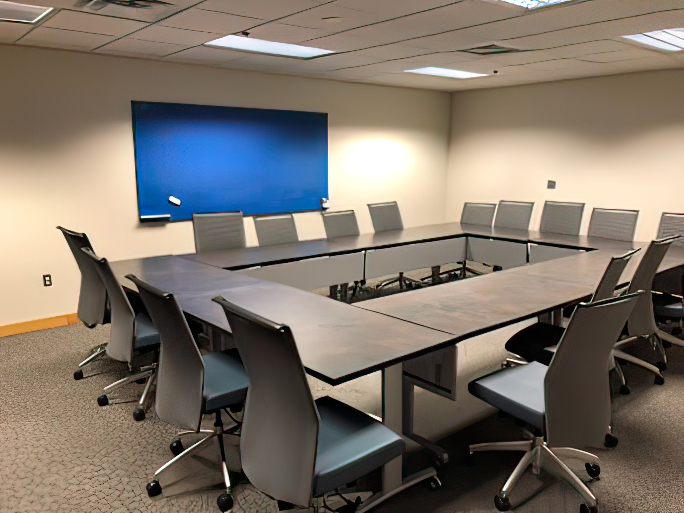An empty room with a large table surrounded by chairs. A marker board hangs on the wall nearby.