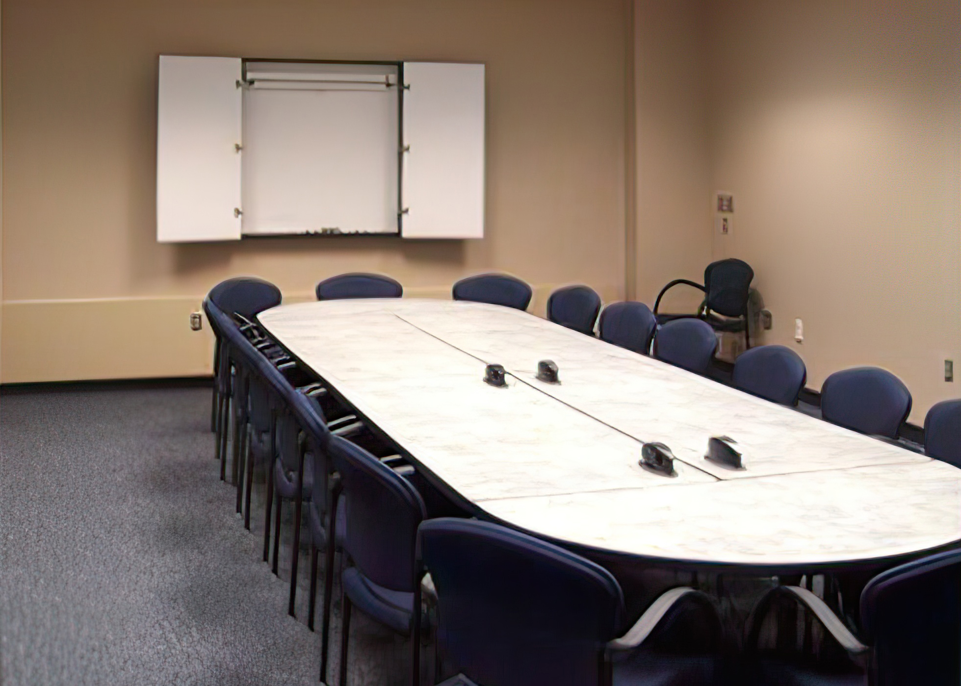 A long, conference table, surrounded by chairs, faces a wall with an open cabinet with a whiteboard.
