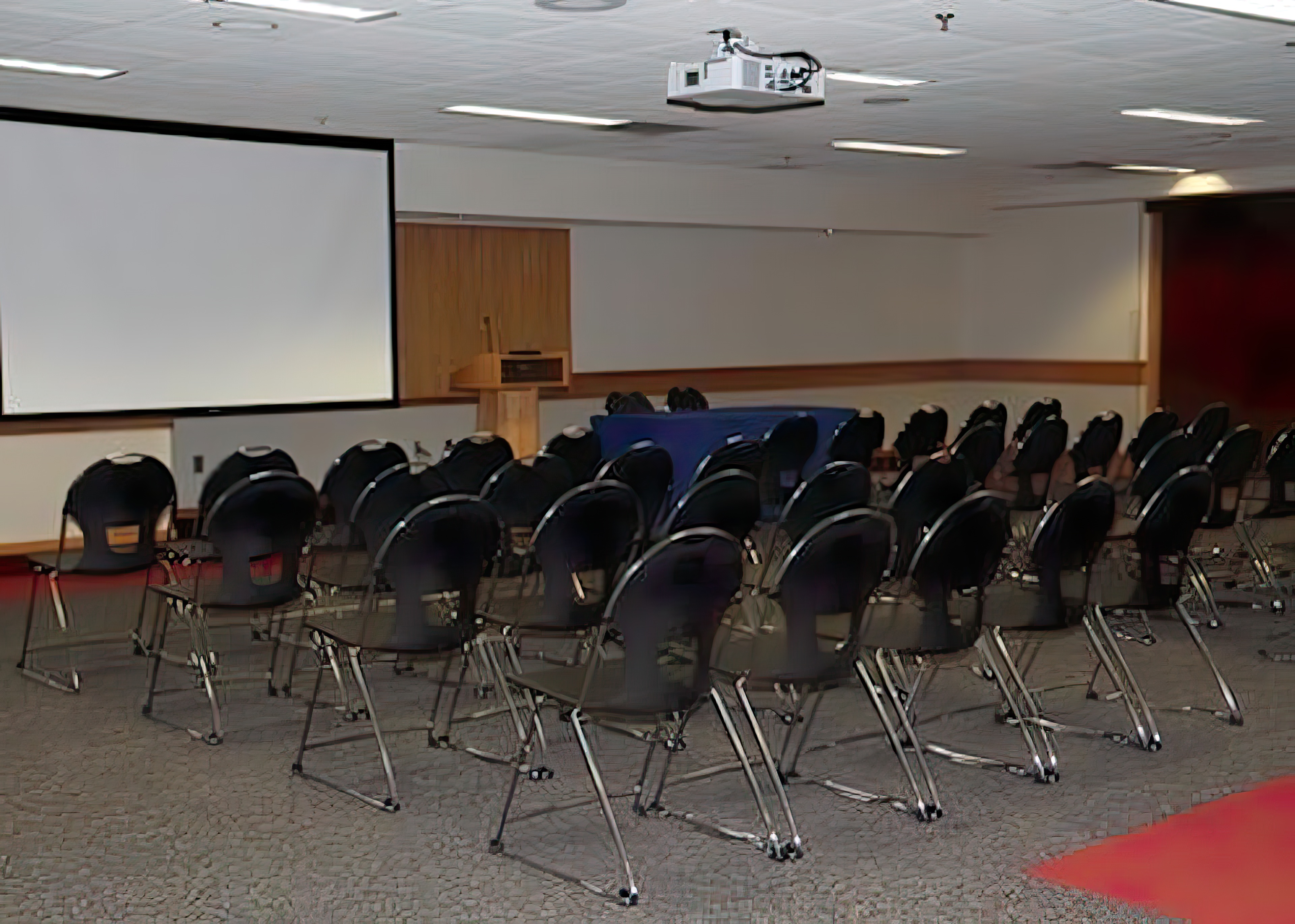 Rows of chairs face a speakers podium. A view screen has been pulled down in the background.