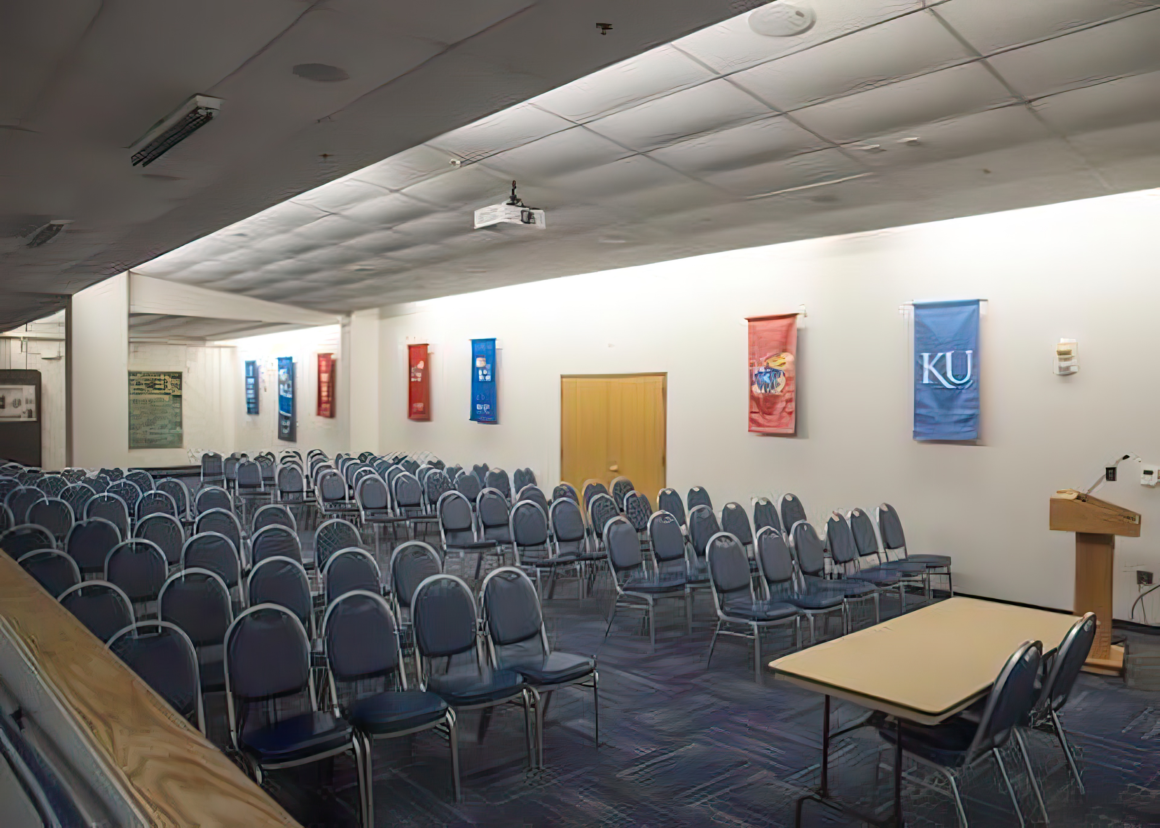 A room set up with chairs in rows, a table and podium in front, and KU banners hanging on the wall. 