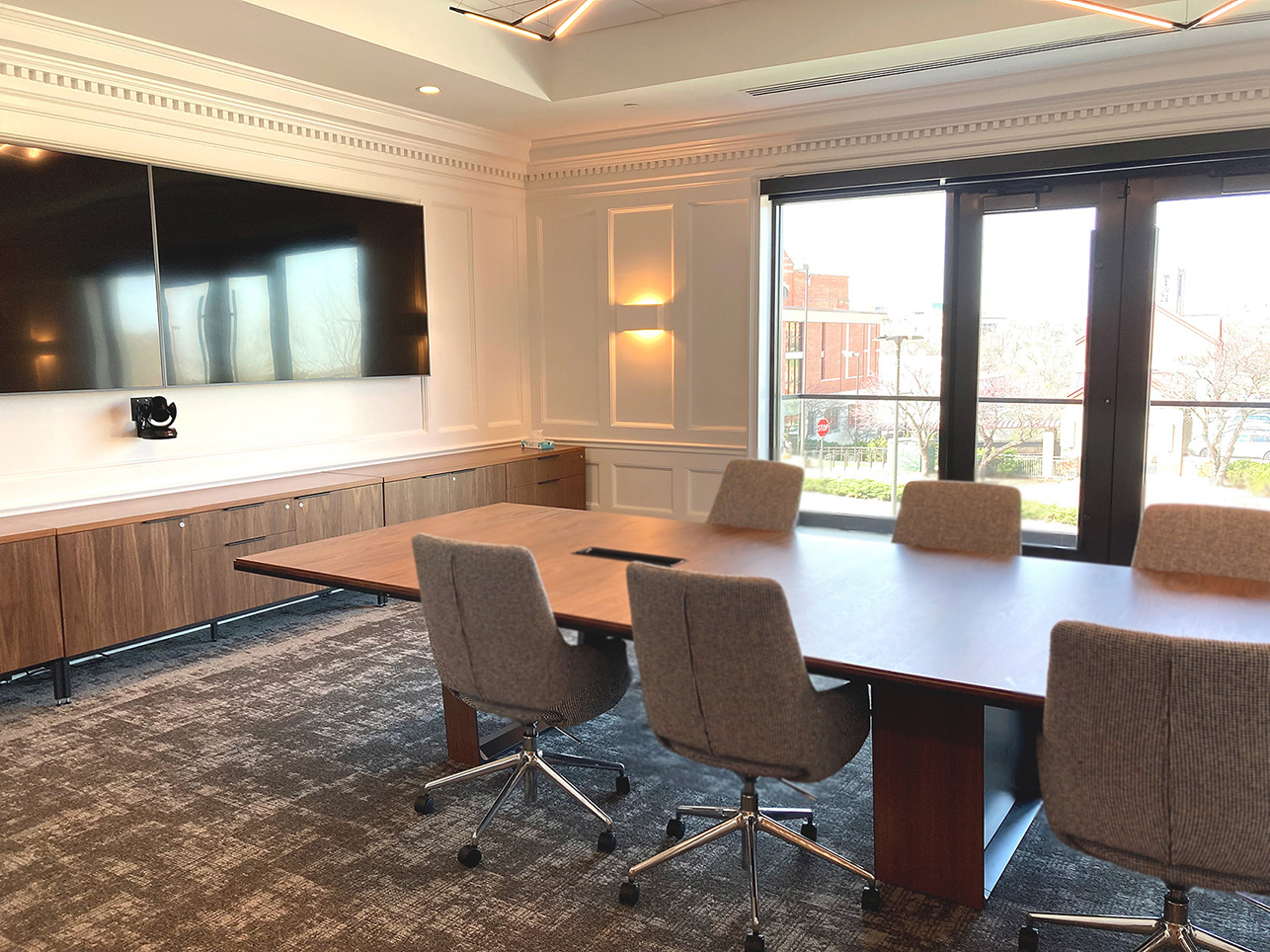 A long conference table, surrounded by office chairs on two sides, face two video monitors.