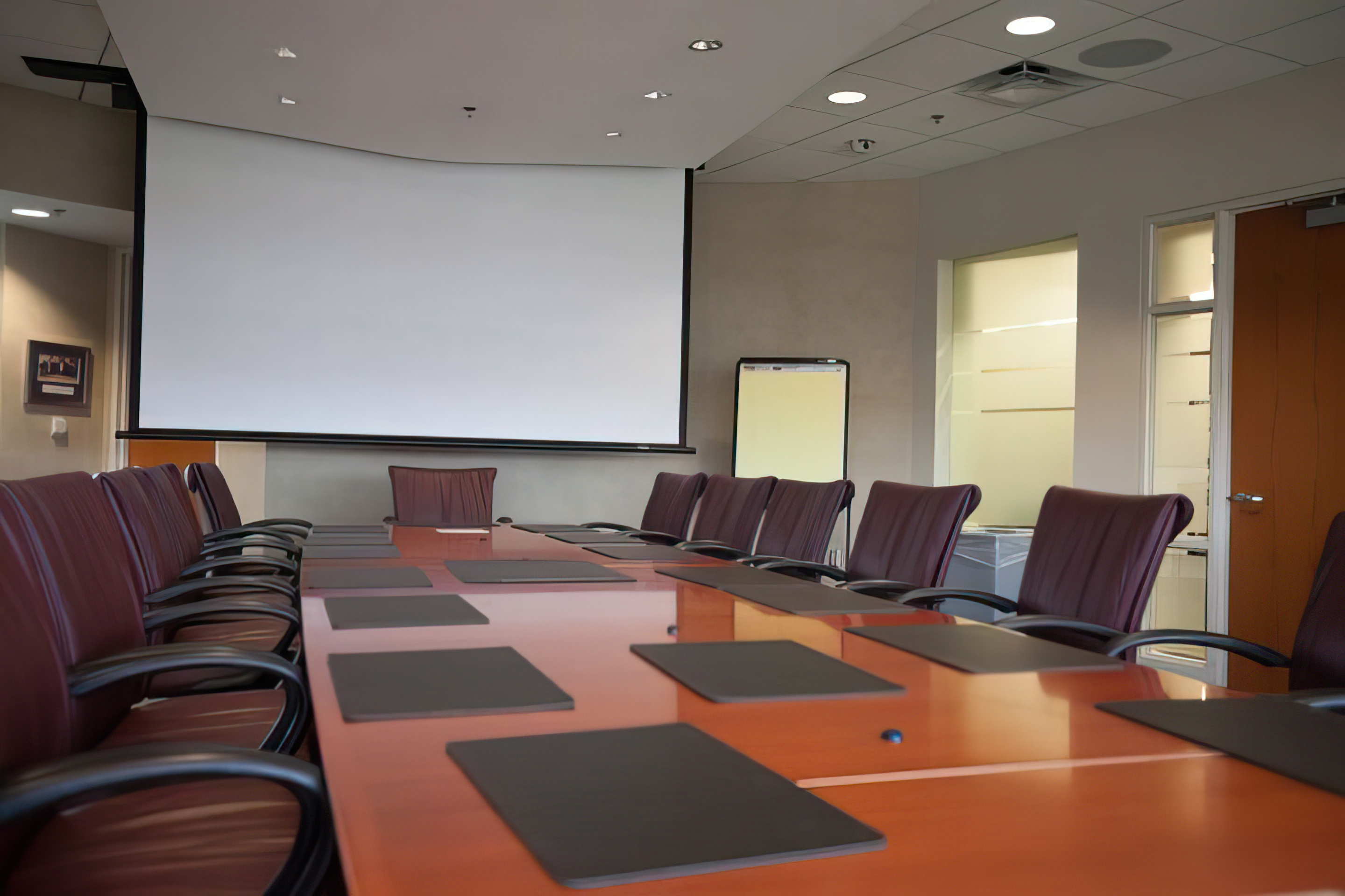 A large conference table, surrounded by office chairs, faces a pull-down projection screen.
